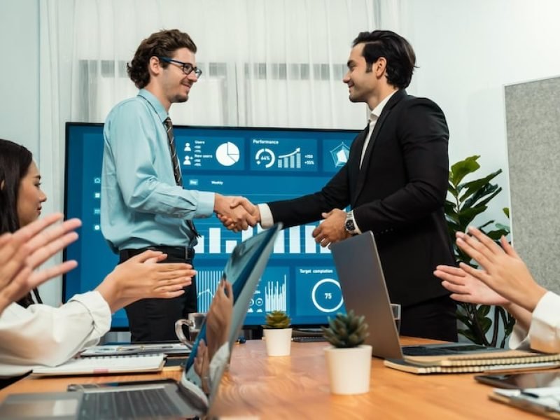 two-businessman-shake-hand-meeting-room-with-cheerful-colleagues-habiliment_31965-685689